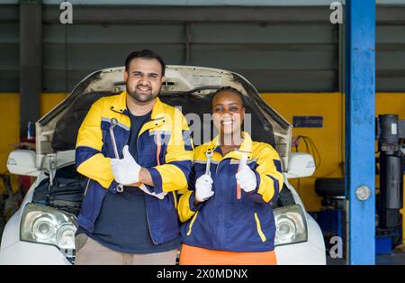 Zwei Mechaniker in Uniform und Schutzhandschuh heben die Daumen nach oben, stehen mit geöffneter Motorhaube vor einem Fahrzeug und lächeln, während sie den Schraubenschlüssel halten. Stockfoto