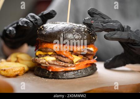 Weibliche Hände in schwarzen Handschuhen halten einen großen schwarzen Burger mit Rindfleisch und verzehrfertig, selektiv fokussiert. Stockfoto