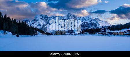 Panoramablick auf den gefrorenen und schneebedeckten See Lago di Misurina mit der Berggruppe Punta Sorapiss in der Ferne im Winter bei Sonnenuntergang. Stockfoto