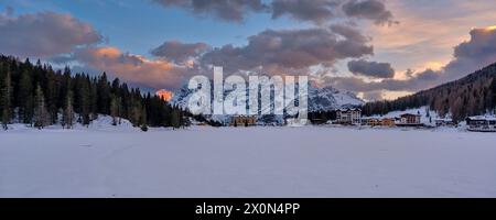 Panoramablick auf den gefrorenen und schneebedeckten See Lago di Misurina mit der Berggruppe Punta Sorapiss in der Ferne im Winter bei Sonnenuntergang. Stockfoto