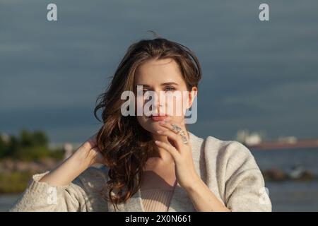 Fröhliche brünette Frau mit frischer Haut, natürlichem Make-up und gesundem, welligen braunen Haar genießt die sonnige Morgen- und Meeresluft. Weibliches Outdoormodell Stockfoto