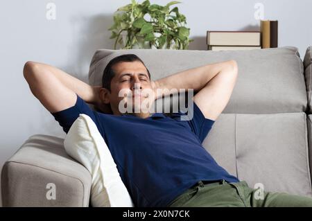 Glücklicher Mann, der sich mit Armen auf dem Kopf entspannt und in der Nacht zu Hause auf einer Couch sitzt Stockfoto