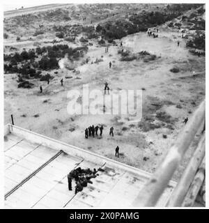 Deutsche Soldaten einer Landeeinheit ergeben sich, einige Spielkarten auf einer Terrasse und andere kochen am Strand, von einem Dach des Gesundheitszentrums aus gesehen. Stockfoto