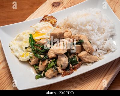 Genießen Sie die Aromen von thailändischem Basilikum-Huhn, serviert mit einem perfekt Spiegelei, Brokkolini und duftendem Jasminreis, elegant präsentiert auf einem weiß Stockfoto