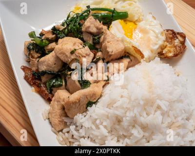 Genießen Sie die Aromen von thailändischem Basilikum-Huhn, serviert mit einem perfekt Spiegelei, Brokkolini und duftendem Jasminreis, elegant präsentiert auf einem weiß Stockfoto