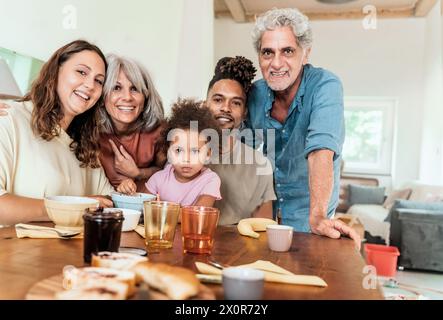 Vielfältige, generationenübergreifende Familien versammeln sich um den Küchentisch und teilen einen freudigen Moment - Großeltern, Eltern und ein Kind lächeln herzlich Stockfoto
