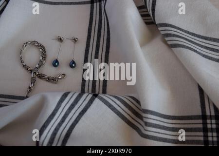 Silberne Ohrringe mit schwarzen Perlen und silbernem Armband auf der weißen Kleidung aus Baumwolle mit sanfter Textur. Käfigtextiler Hintergrund am sonnigen Sommertag Stockfoto