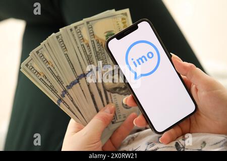 KIEW, UKRAINE - 1. APRIL 2024 IMO-Messenger-Symbol auf Smartphone-Bildschirm und Geld in weiblicher Hand. IPhone-Display mit App-Logo und Hundert-Dollar-Scheine in Mädchenhänden aus nächster Nähe Stockfoto