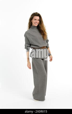 Eine Frau mit einem strahlenden Lächeln, die einen stylischen grauen Pullover und eine Hose mit weitem Bein trägt. Ihr Haar kaskadierte ihren Hals, die Ärmel hochgerollt, verströmt Vertrautes Stockfoto