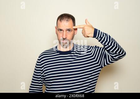 Hispanischer Mann mit Bart in seinen 40ern, der einen gestreiften Pullover trägt, der über einem beigen Hintergrund steht und sich erschoss und tötete, indem er mit der Hand zeigt und Stockfoto