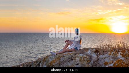 Ein Reisender sitzt auf einem Berggipfel am Meer und schaut auf den Sonnenuntergang. Stockfoto