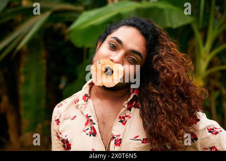 Südasiatischer Mann mit Blume im Mund posiert ausdrucksstarkes Make-up, kühne Weiblichkeit. Das Tragen eines Blumenhemdes in grünem Laub repräsentiert das Geschlecht Stockfoto