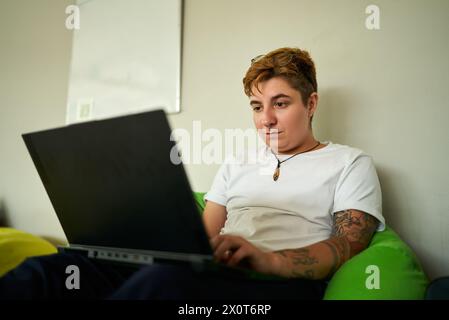 Transgender-Person arbeitet auf einem Laptop, sitzt auf einer bunten Sitztasche in einem entspannten Bürobereich. Konzentriert auf den Bildschirm in legeren Kleidern und Tätowierungen sichtbar Stockfoto