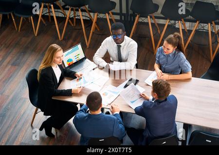 Weibliche CEO leitet Geschäftstreffen und analysiert Diagramme auf Laptops in modernen Büros. Das multirassische Team bespricht die Strategie am Tisch mit Papieren und Diagrammen Stockfoto