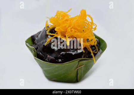 Nahaufnahme eines „umgedrehten“ thailändischen Khanom Tako-Desserts mit schwarzem klebrigem Reis und süßen goldenen Eigelb-Fetzen (Foi Thong) in einer Pandan-Blatttasse Stockfoto