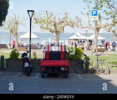 APE-Fahrzeug auf dem Parkplatz mit ungewöhnlichen Bäumen und Marktständen in Marta mit Blick auf den Bolsenasee, Provinz Viterbo, Region Latium, Italien. April 2024 Stockfoto