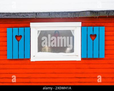 Die bezaubernde rote Holzfassade eines typisch schwedischen Hauses ist mit leuchtend blauen Fensterläden mit herzförmigen Ausschnitten geschmückt. Der Schnee liegt sanft auf Stockfoto