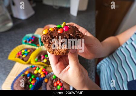 Kinderhand hält hausgemachte Schokoladen-Cupcakes mit Schokoladenchips und bunten Süßigkeiten. Stockfoto