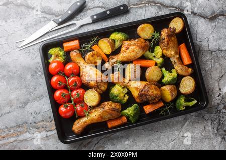 Im Ofen gebackene Hähnchenkeulen mit frisch gebackenem Gemüse in Nahaufnahme auf einem Backblech auf dem Tisch. Horizontale Draufsicht von oben Stockfoto