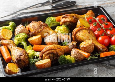 Leckere gebackene, ruddy Hähnchenschenkel mit Kartoffeln, Brokkoli, Tomaten, Karotten, Nahaufnahme auf einem Backblech auf dem Tisch. Horizontal Stockfoto