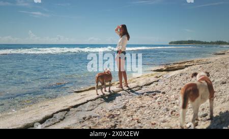 Eine Frau steht an einem Sandstrand neben zwei Hunden. Die Frau blickt auf den Ozean, während Hunde im Sand spielen. Die Szene fängt einen friedlichen Moment der Gemeinschaft zwischen Mensch und Tier ein. Stockfoto