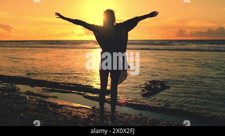 Frau hebt die Hände auf dem Sonnenuntergang Sandstrand, Ozeanwellen im Hintergrund. Weibliche Silhouette steht an der Küste des Ozeans. Der orangefarbene Himmel spiegelt sich im Wasser. Entspannen Sie sich, genießen Sie den Sommerurlaub. Rückansicht Stockfoto