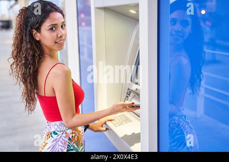 Eine junge Frau, die mit ihrem Smartphone Bargeld an einem Geldautomaten abhebt und in die Kamera schaut Stockfoto