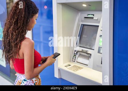 Eine junge Frau, die mit ihrem Smartphone Bargeld an einem Geldautomaten abhebt Stockfoto