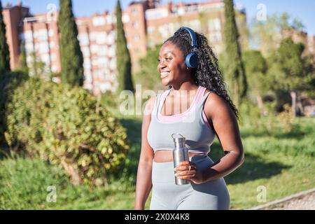 Bild einer weiblichen 20er-Frau, die Kopfhörer trägt und eine Wasserflasche hält, während sie durch den grünen Park geht. Kurvige afrikanerin während der Trainingsroutine Outd Stockfoto