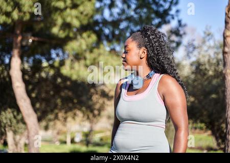Eine große afroamerikanische Frau, die an einem Sommertag während des Spaziergangs im Park Handy und Kopfhörer benutzt Stockfoto