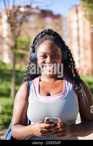 Vertikales Porträt einer jungen afroamerikanischen Frau, die Musik mit Kopfhörern hört, während sie ein Smartphone hält und in die Kamera schaut. Stockfoto