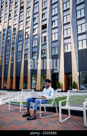 Junger afroamerikanischer Geschäftsmann, der auf der Bank in der Stadtstraße sitzt. Lebensstil beruflicher männlicher Arbeit im städtischen Umfeld. Stockfoto