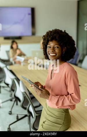 Ccheerful Afroamerikanerin leitet ein Meeting und hält sich an einem digitalen Tablet fest, während Kollegen hinter ihr Gespräche führen. Stockfoto