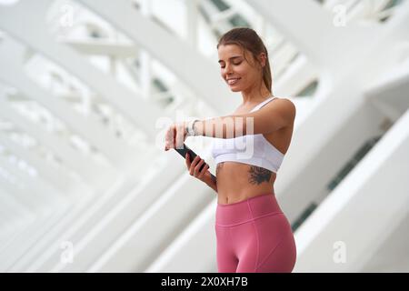 Frau in aktiver Hose lächelt, überprüft die Zeit in der Wache, während sie das Handy hält Stockfoto