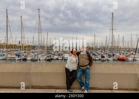 Lächelnde Touristen stehen an der Promenade von Puerto Vell, zahlreiche Segelboote vor Anker am Pier im Hintergrund, ruhiger Nachmittag in der Stadt mit bewölktem Wetter Stockfoto