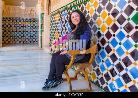 Lateinamerikanische Frau mittleren Alters, die auf Holzstuhl im Flur mit Mosaikwänden im Palast sitzt, auf die Kamera blickt, dunkle, bunte Kleidung, Touris Stockfoto