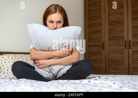 Einsame Frau sitzt auf dem Bett im Schlafzimmer und umarmt Kissen. Depressive Frau mit leerem Ausdruck. Stockfoto