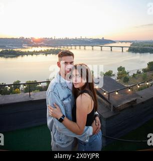 Ein Paar teilt einen zärtlichen Moment auf einem Dach, seine Silhouetten sind gegen den Abendhimmel geätzt. Das Leuchten der Stadt verstärkt die romantische Atmosphäre. Stockfoto