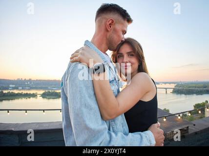 Ein junges Paar genießt einen ruhigen Abend auf einem Dach, umgeben von den warmen Farbtönen der untergehenden Sonne. Ihre Umarmung spricht von gemeinsamen Momenten und Ruhe Stockfoto