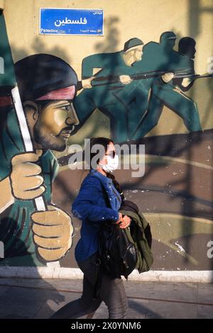 Teheran, Iran. April 2024. Eine iranische junge Frau, die eine Gesichtsmaske trägt, ohne ihr obligatorisches islamisches Kopftuch zu bedecken, geht neben einem Wandgemälde von Soldaten der iranischen Revolutionsgarde (IRGC) auf dem Palästinensischen Platz in der Innenstadt von Teheran. Israel feierte am Sonntag seine Luftverteidigung angesichts eines beispiellosen Angriffs des Iran und sagte, dass die Systeme 99 % der mehr als 300 Drohnen und Raketen, die auf sein Territorium abgefeuert wurden, vereitelten. (Kreditbild: © Rouzbeh Fouladi/ZUMA Press Wire) NUR REDAKTIONELLE VERWENDUNG! Nicht für kommerzielle ZWECKE! Stockfoto