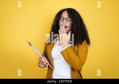 afroamerikanische Geschäftsfrau mit Papierkram in den Händen über gelbem Hintergrund gelangweilt Gähnen müde Mund mit Hand bedeckt. Ruhelos und Schläfrigkeit. Stockfoto