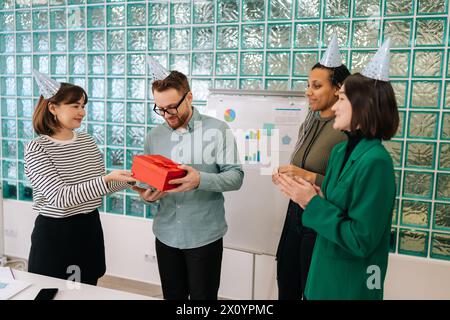 Wide Shot Business-Team von Büroangestellten in festlichen Mützen in modernen Büros als Geschenk für gutaussehenden männlichen Kollegen zum Geburtstag. Fröhlicher Mann überrascht Stockfoto