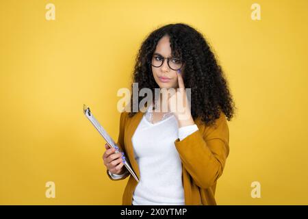 afroamerikanische Geschäftsfrau mit Papierkram in den Händen über gelbem Hintergrund, der auf das Auge zeigt, wie Sie eine Geste sehen, misstrauischer Ausdruck Stockfoto