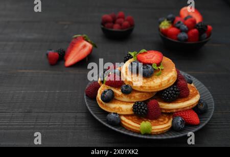 Flache Lage von gestapelten goldenen Pfannkuchen auf schwarzem Teller mit frischen Beeren im dunklen Stil auf schwarzem hölzernem Hintergrund Stockfoto