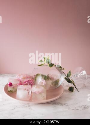 Schöne kleine rosa Blumen in Eiswürfeln und Glas mit Blütenblättern auf rosa Platte auf rosa Hintergrund. Stockfoto