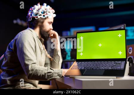 Mann mit EEG-Headset, das Code schreibt, um den Geist in die virtuelle Welt zu transferieren, Laptop mit isoliertem Bildschirm auf dem Schreibtisch Transhumanist mit neurowissenschaftlicher Technologie und Chroma Key Notebook, um Superintelligenz zu gewinnen Stockfoto