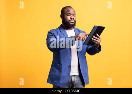 Gelangweilte Person, die auf dem Tablet-Bildschirm gescrollt ist, isoliert über Studiohintergrund. afroamerikanischer Mann, der auf einem tragbaren Gerät Zeit in sozialen Medien verbringt und Freizeit genießt Stockfoto