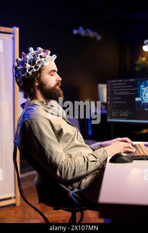 Mann mit EEG-Headset, der auf der Tastatur schreibt und Code schreibt, sodass er den Geist in die virtuelle Welt des Computers transferieren kann. Transhumanist schließt die Augen, transzendiert, wird eins mit KI Stockfoto