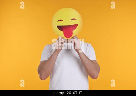 Mann, der das Gesicht mit Emoticon bedeckt, der die Zunge auf gelbem Hintergrund hinausragt Stockfoto