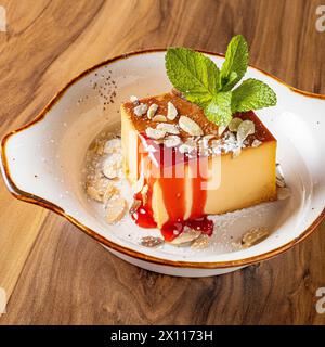 Flan-Dessert serviert auf einem schönen Steingut-Teller, weiß garniert mit gerösteten Mandeln, Erdbeersauce und frischer Minze Stockfoto